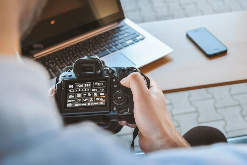 pexels photo 442573 442573 Close-up of hands holding DSLR camera at a workspace with laptop and smartphone.
